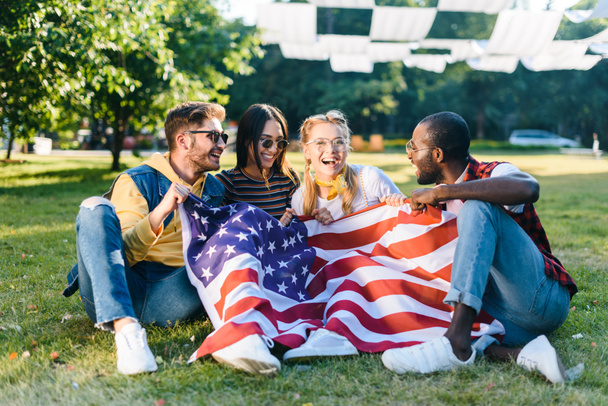 wielorasowe znajomych szczęśliwy z amerykańską flagę siedzi na zielonej trawie w parku - Zdjęcie, obraz