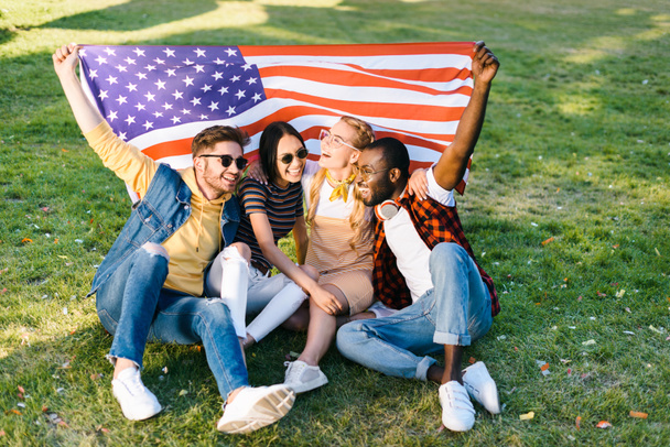 многорасовые веселые друзья с американским флагом, сидящие на зеленой траве в парке
 - Фото, изображение