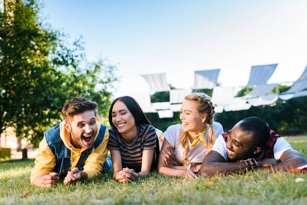 portrait of cheerful interracial young friends lying on green grass in park on summer day - Photo, Image