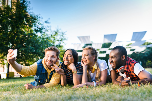 portrait of multiethnic cheerful friends taking selfie on smartphone while resting on green grass in park - Foto, imagen