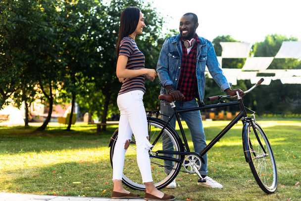χαμογελώντας πολυφυλετικής ζευγάρι με ρετρό ποδήλατο έχοντας συνομιλία στο πάρκο - Φωτογραφία, εικόνα