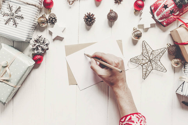 Kind schreibt Brief an Weihnachtsmann von oben. Kinderhände halten Bleistift in der Hand und basteln Wunschliste und Papier mit Weihnachtsschmuck und Ornamenten und Geschenken auf weißem Holz. Raum für Text - Foto, Bild