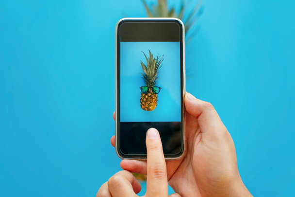 Instagram fotografie ovoce. ruce drží telefon a pořizování fotografií z ananasu v sluneční brýle na modrém papíře, trendy plochý ležela. Stylový food fotografie. Creative telefon obrázek - Fotografie, Obrázek