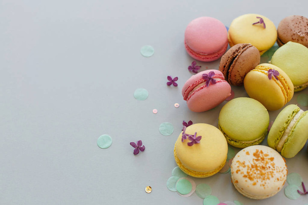 colorful macarons on trendy pastel gray paper with lilac flowers and confetti. tasty pink, yellow, green and brown macaroons. candy bar for party. food photography. yummy background - Foto, imagen