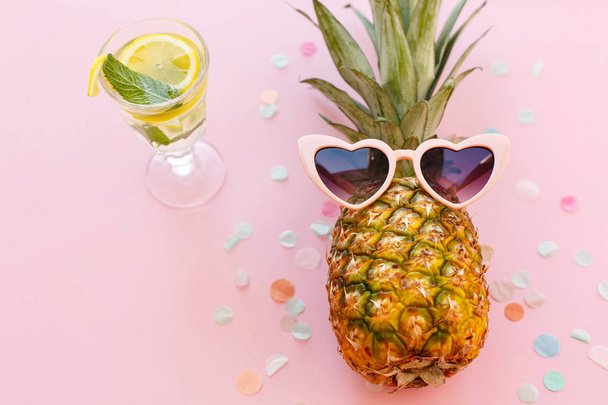 pineapple in pink sunglasses and mojito cocktail drink on trendy pink paper background with confetti. summer vacation and party concept. space for text. summer holidays - Photo, Image