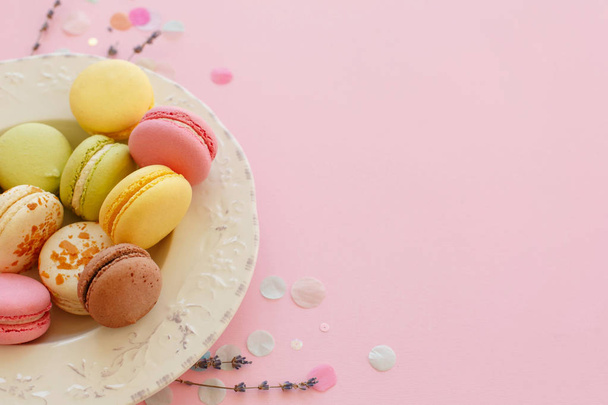 leckere bunte Macarons im Vintage-Teller auf trendigem pastellrosa Papier mit Lavendel und Konfetti. Raum für Text rosa, gelb, grün, weiß, braune Makronen. Moderne Food-Fotografie - Foto, Bild