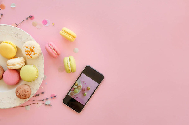 instagram food photography of tasty colorful macarons in plate and phone on trendy pastel pink paper flat lay. space for text. modern photo workshop. holiday catering - Foto, Bild