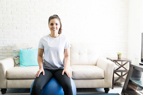 sonriente joven sentada en bola suiza para hacer ejercicio en la sala de estar
 - Foto, imagen