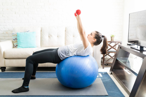 Ajuste mujer hispana joven inclinándose sobre la pelota de estabilidad y haciendo ejercicios de manos con pesas
 - Foto, imagen