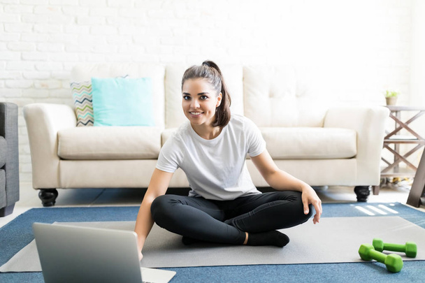 young latin woman sitting on exercise mat at home and watching workout tutorial online on laptop - Foto, immagini