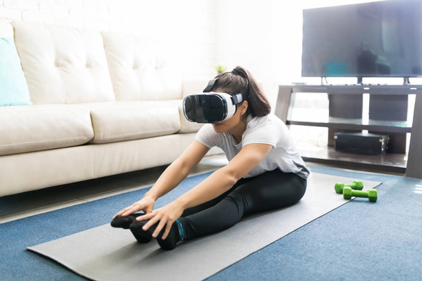 Woman in virtual reality glasses doing stretching workout on exercise mat at home - Foto, afbeelding