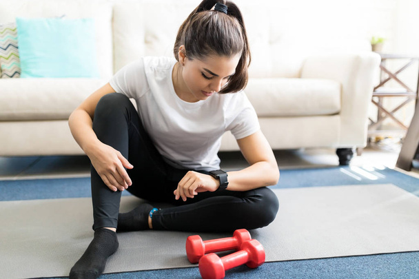 young woman sitting on yoga mat in living room and setting smartwatch before workout - Photo, Image