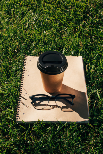 close up view of coffee to go, notebook and eyeglasses on green grass - Фото, изображение