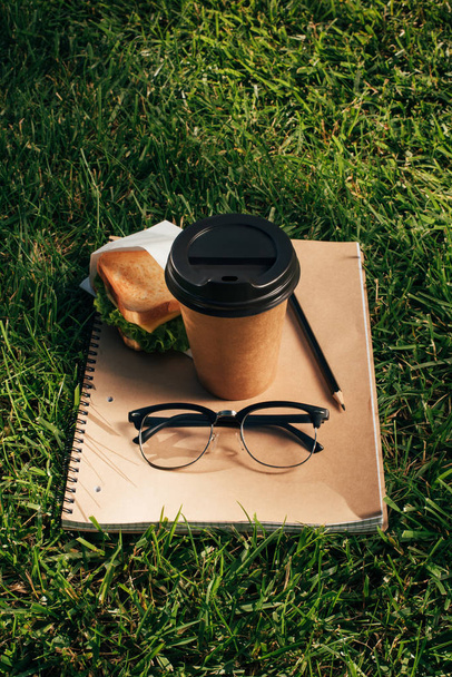 close up view of notebook, sandwich, eyeglasses and coffee to go on green grass - 写真・画像