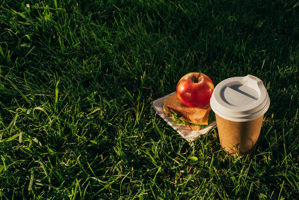 Close-up van koffie te gaan, sandwich en apple op groen gras - Foto, afbeelding