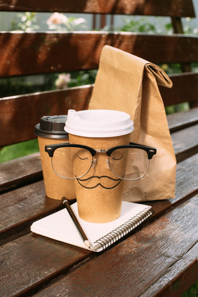 close up view of disposable cups of coffee, paper bag, eyeglasses and notebook on bench - Fotoğraf, Görsel
