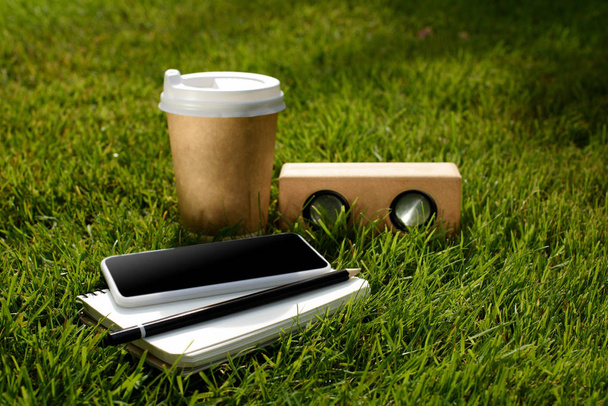 close up view of coffee to go, smartphone, notebook and audio speaker on green grass - Fotografie, Obrázek