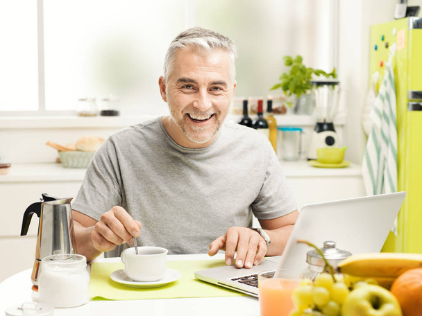 Smiling mature man having an healthy breakfast at home, he is sitting at the kitchen table, drinking coffee and connecting with his laptop - Zdjęcie, obraz