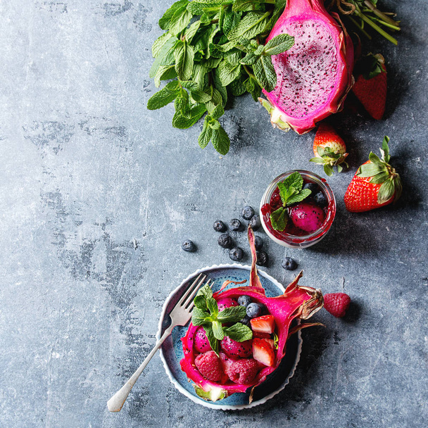 Vegan fruit salad with berries and mint served in pink dragon fruit with ingredients above on ceramic plate over blue texture background. Top view, space. Healthy eating - Foto, Bild