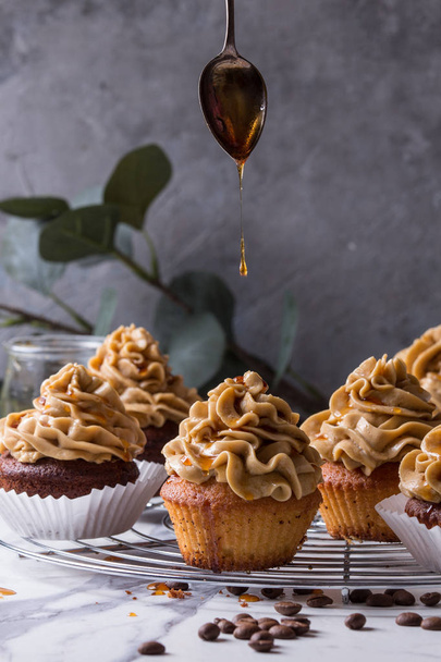 Fresh baked homemade cupcakes with coffee buttercream and pouring from spoon caramel standing on cooling rack with eucalyptus branch and coffee beans above over white marble kitchen table. - Foto, imagen