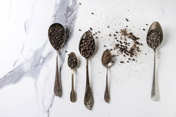 Variety of different black peppers allspice, pimento, long pepper, monks pepper, peppercorns and ground powder in vintage spoons over white marble texture background. Top view, space. - Fotoğraf, Görsel