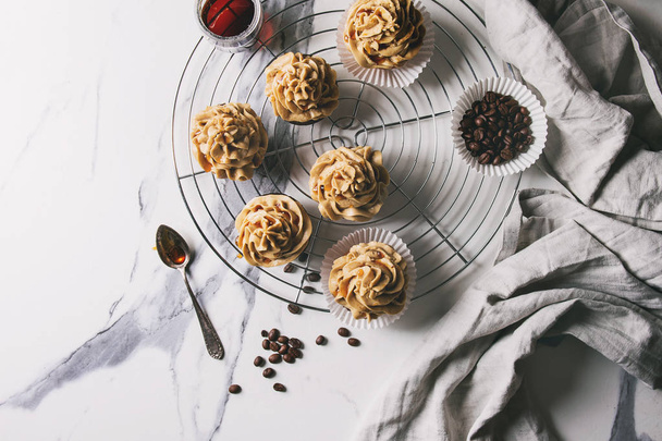 Fresh baked homemade cupcakes with coffee buttercream and caramel standing on cooling rack with coffee beans above over white marble background. Flat lay, space - 写真・画像