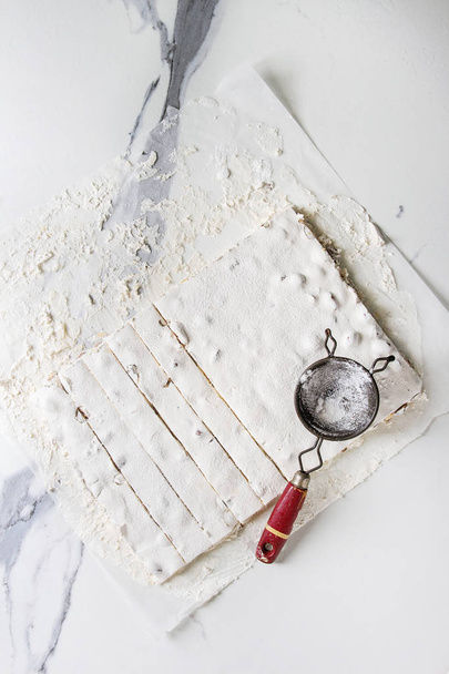 Homemade honey nuts nougat turron sliced on crumpled paper with vintage sieve and sugar powder over white marble background. Flat lay, space. - Foto, Imagem