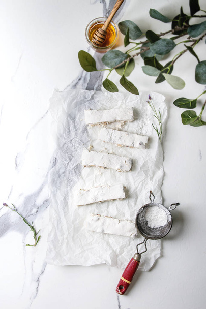 Homemade honey nuts nougat turron sliced on crumpled paper with lavender, glass of honey and sugar powder over white marble background. Flat lay, space. - Photo, image