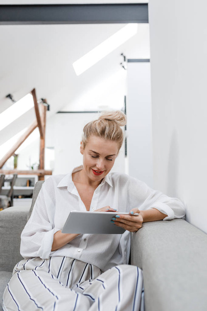 Pretty blonde smiling Caucasian woman sitting on couch at home and typing on tablet. - Foto, Bild