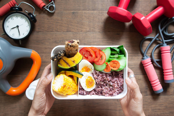 Frauenhände halten Lunchbox mit Reisbeeren, gekochten Eiern, Zuckermais, Kürbis, Tomaten und Müsliriegeln, Draufsicht mit Sport- und Fitnessgeräten auf Holzgrund - Foto, Bild