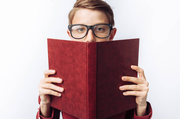 Portrait of a positive and emotional schoolboy looking out from behind a book, white background, glasses, red shirt, business theme, - Zdjęcie, obraz