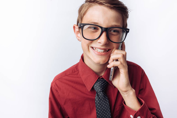 Portrait of positive and emotional schoolboy talking on phone, white background, glasses, red shirt, business theme, - Fotografie, Obrázek
