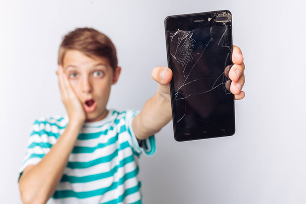 Portrait of a beautiful and emotional boy, in whose hands a broken phone shows surprise, white background, blue t-shirt, - Фото, изображение