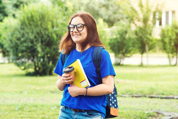 Молодой улыбающийся студент ходит с книгами, рюкзаком и кофе, чтобы пойти, в летний день
 - Фото, изображение