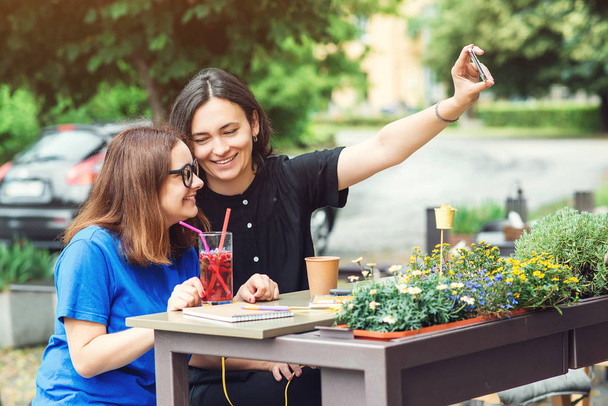Due ragazze sorridenti che si fanno un selfie sullo smartphone, sedute alla caffetteria all'aperto, fanno una pausa insieme in una giornata estiva
. - Foto, immagini
