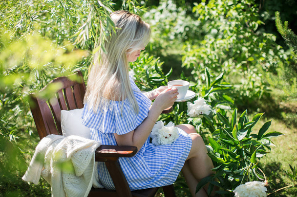 young woman is resting in the garden on a chair with a mug of tea - Фото, изображение