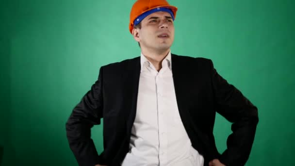An attractive man in a orange construction helmet and suit looks at the camera. A young worker shows gestures with his hands on green chroma key background - Filmagem, Vídeo
