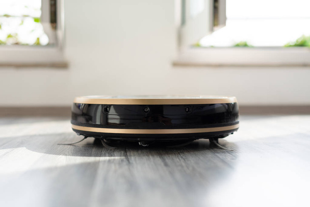aspiradora robótica en suelo de madera laminada con ventanas brillantes en el fondo, tecnología de limpieza inteligente moderna y conveniente
 - Foto, Imagen