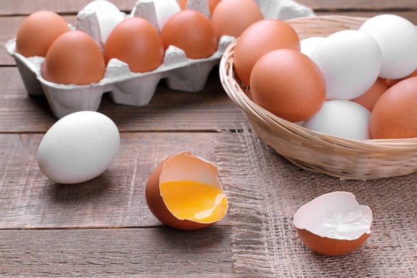 Chopped chicken egg with yolk close-up next to eggs in basket and tray on brown wood background - Photo, Image