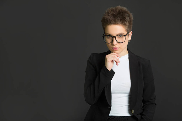 beautiful young girl with short hair, in a black suit and wearing glasses on a black background with a serious face - Photo, image