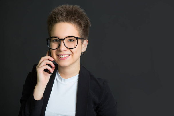 beautiful young girl with short hair, in a black suit and wearing glasses on a black background with a phone in her hand - Photo, image