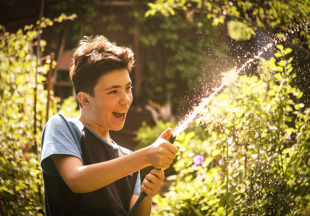 kid hands hold hose with squirting water on the summer sunny green garden - 写真・画像