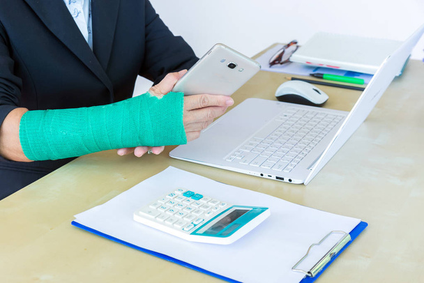 femme d'affaires avec coulée verte sur le bras tenant le téléphone intelligent et travaillant sur un ordinateur portable au bureau, se concentrer sur la main cassée
. - Photo, image