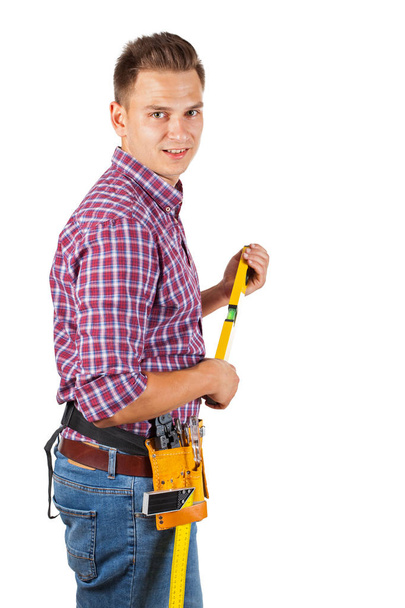 Aantrekkelijke jonge reparateur met gele helm en gereedschap gordel op geïsoleerde achtergrond - Foto, afbeelding