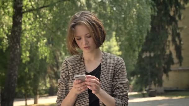 Young attractive concentrated girl is typing message on smartphone in park in daytime, in summer, communication concept. - Filmati, video