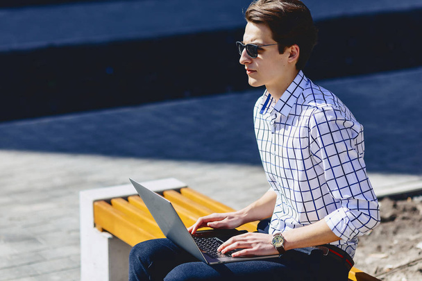 Young stylish man with phone work on laptop at park on sunny day - Valokuva, kuva