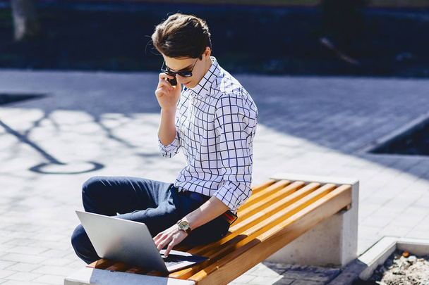 Молодий стильний чоловік з телефоном працює на ноутбуці в парку в сонячний день
 - Фото, зображення