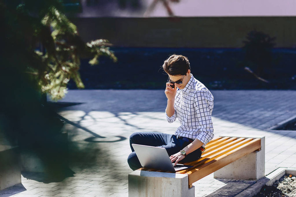 Молодой стильный мужчина с телефоном работает на ноутбуке в парке в солнечный день
 - Фото, изображение