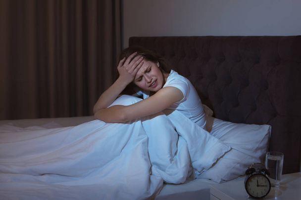 Νεαρή γυναίκα με τρομερό πονοκέφαλο που κάθεται στο κρεβάτι τη νύχτα - Φωτογραφία, εικόνα