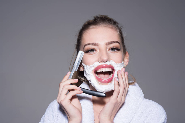 Cheerful blond woman in white bathrobe having fun in bathroom isolated on gray background. Lovely female applying shaving foam on face and holding cut-throat razor, femininity and masculinity concept - 写真・画像